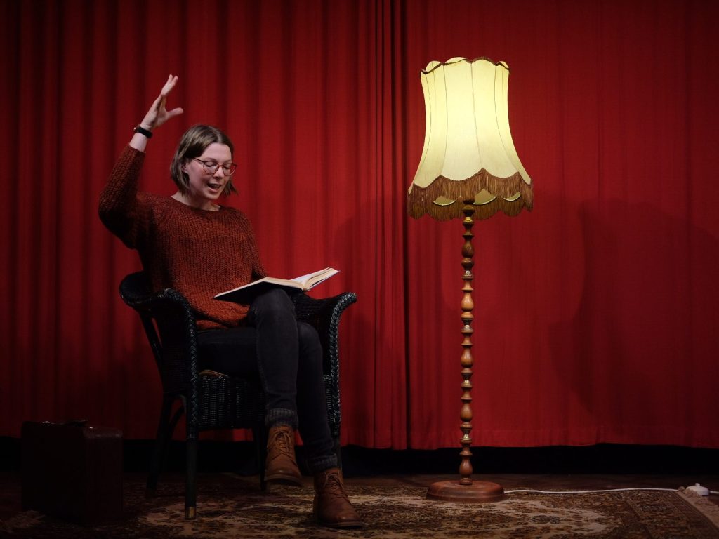 Foto: Lena sitzt vor einem roten Vorhang neben einer alten Stehlampe und liest aus einem Buch vor.